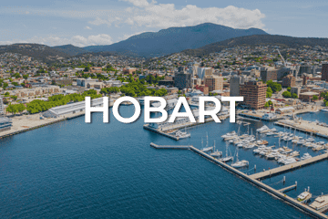 Aerial view of Hobart's harbor with sailboats docked along the marina. The city skyline, a thriving hub with numerous businesses for sale, is set against green hills and a mountain backdrop under a partly cloudy sky. The word 'HOBART' is prominently displayed over the image.